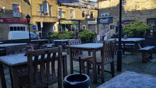 Outdoor seating area with wooden tables and chairs, a coffee cup in the foreground, and stone buildings in the background.