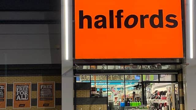 Brightly lit Halfords store entrance at night, featuring large orange signage and a display of various products outside.