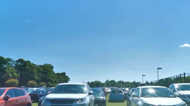 Busy car park filled with various vehicles under a clear blue sky, surrounded by greenery.