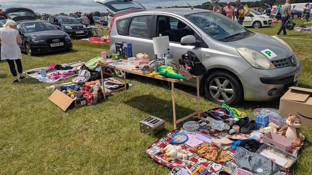 Car boot sale with various items displayed on tables, parked cars in the background, and people browsing in a grassy field.