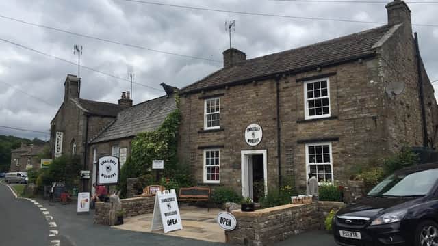 Stone shopfront with "Swaledale Woollens" sign, potted plants, open sign, and parked cars on a cloudy day in Muker.