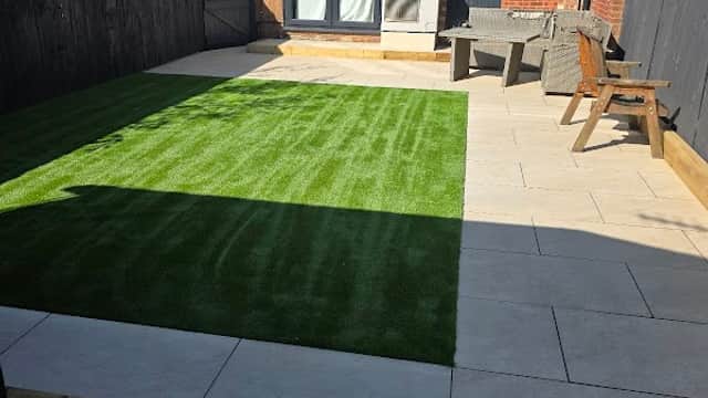 Lush green artificial grass area with tiled patio, wooden chairs, and a wooden fence in a sunny garden setting.