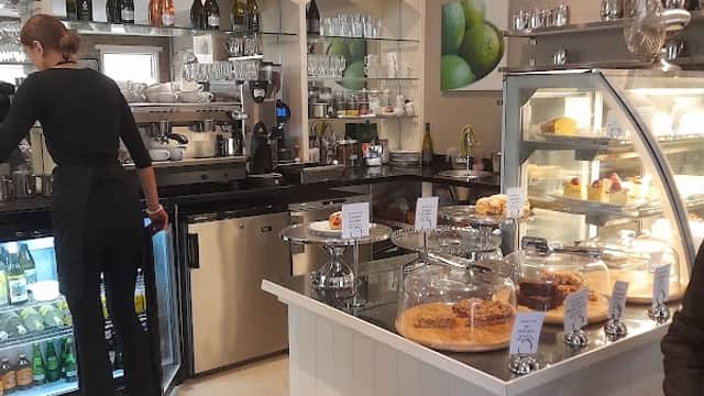 Cozy cafe interior with a counter displaying cakes, drinks, and a barista preparing coffee. Green lime decor visible.