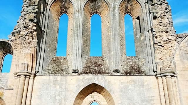 Ruins of a historic stone building with large arched windows under a bright blue sky, surrounded by green grass.