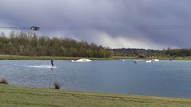 A person water skiing on a lake with a cable system, surrounded by greenery and dark clouds in the background.