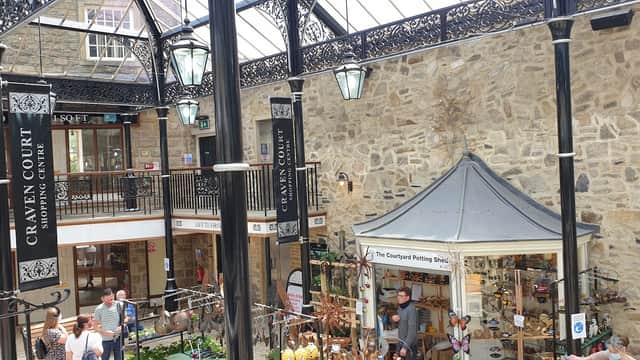 Indoor market featuring stone walls, decorative ironwork, stalls with crafts, and shoppers browsing.