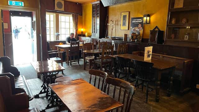 Cozy pub interior with wooden tables, chairs, and warm yellow walls; a lone patron sits at a table near the entrance.