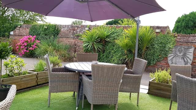 Outdoor seating area with wicker chairs, a round table, and vibrant flowers against a rustic brick wall.