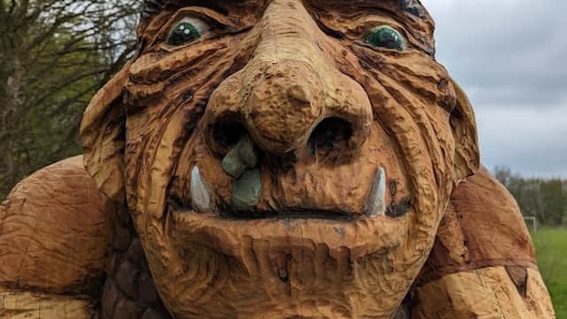 Wooden sculpture of a troll with exaggerated features, set against a backdrop of trees and cloudy skies in Oxley Park.