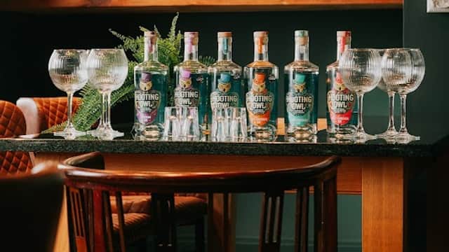 A table with various bottles of Hooting Owl gin, crystal glasses, and copper distilling equipment in a cozy distillery setting.