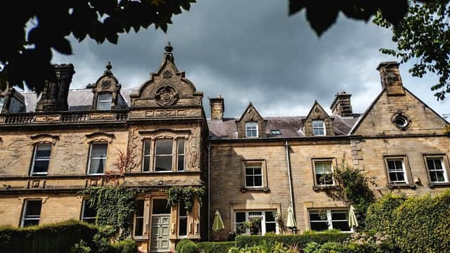 Victorian-style manor with ornate stonework, lush greenery, and outdoor seating under umbrellas against a cloudy sky.