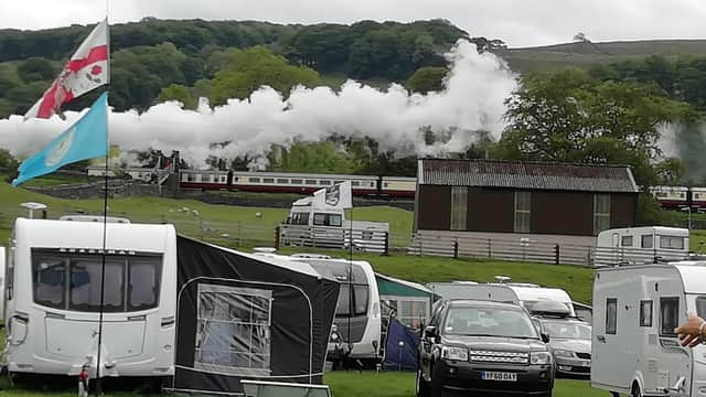 Steam train chugs through green landscape with caravans and tents in the foreground, set in Langcliffe, Yorkshire.