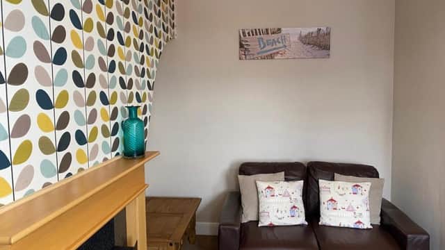 Cozy living room with patterned leaf wallpaper, brown leather sofa, wooden fireplace, and a teal vase on a wooden table.