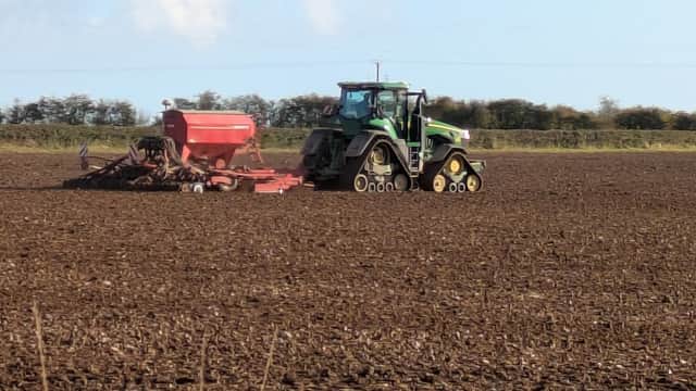 A green tractor with a red seed drill is working in a plowed field under a clear blue sky.