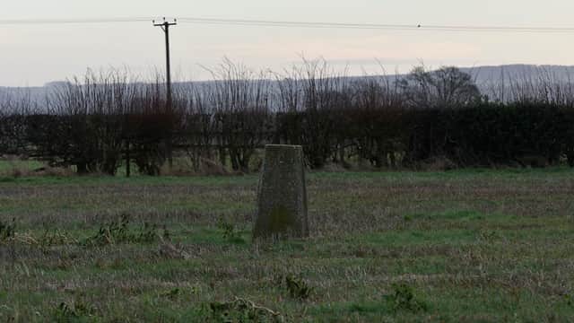 Trig point on a grassy field, surrounded by hedgerows and distant hills under a soft sky.