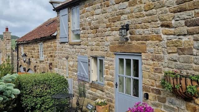 Charming stone cottage with blue shutters, potted flowers, and a small outdoor seating area on a grassy hillside.