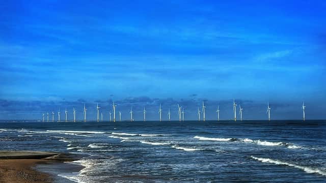 Waves gently lapping at the shore, with a line of wind turbines visible on the horizon under a blue sky.