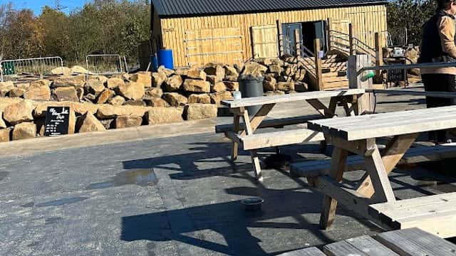 Wooden farm building with a black roof, surrounded by large rocks and picnic tables under a clear blue sky.