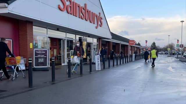 Sainsbury's storefront with shoppers and trolleys outside, cloudy sky, and parking area visible.