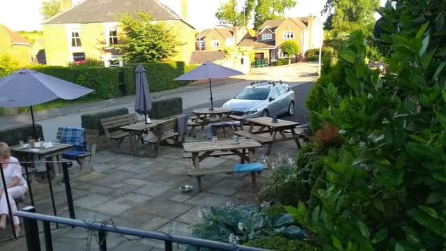 Outdoor seating area with wooden tables and umbrellas, surrounded by greenery, near a quiet street in Little Weighton.