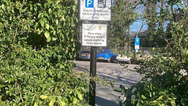 Electric vehicle charging point sign surrounded by greenery, with a view of a road and parked cars in the background.