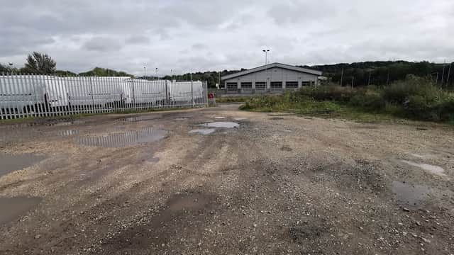 Gravel car park with puddles, surrounded by greenery and a building in the background, near Centenary Riverside Nature Park.