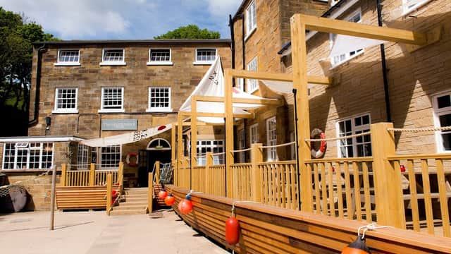 Stone building with wooden deck, lifebuoys, and white awnings under a blue sky in a coastal setting.