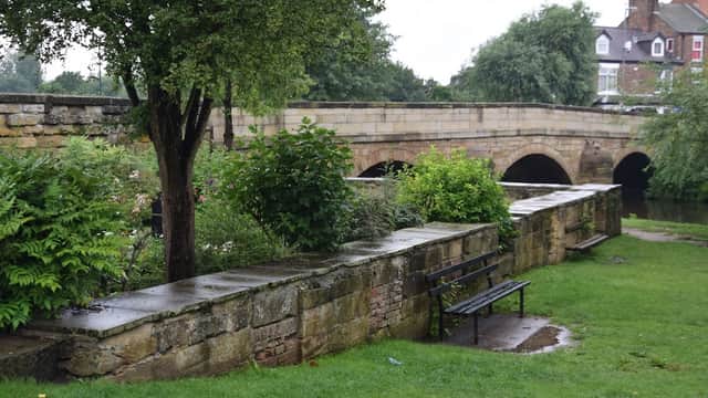 Stone wall with a bench, lush greenery, and a bridge over a river in a serene garden setting.
