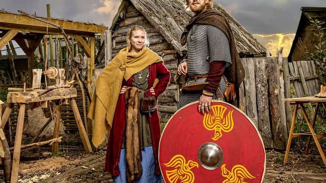 Two people in Viking attire stand by a wooden hut with a red shield, surrounded by rustic tools and a dramatic sky.