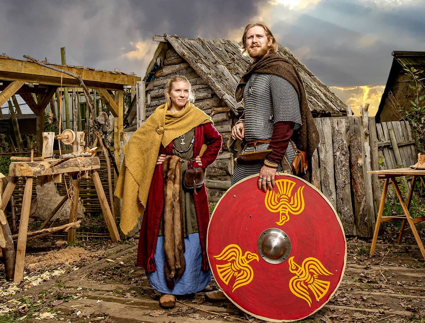 Two people in Viking attire stand by a wooden hut with a red shield, surrounded by rustic tools and a dramatic sky.