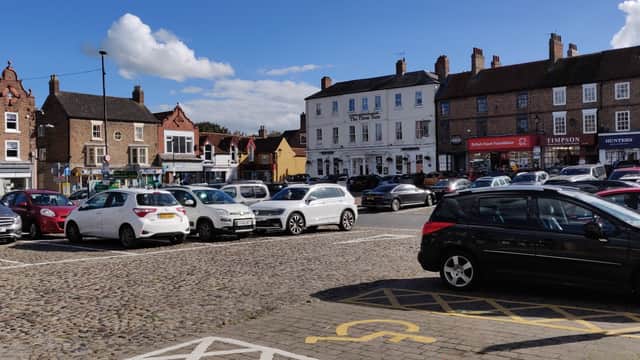 Bus stop area in Thirsk with a busy parking lot, shops, and historic buildings under a blue sky with clouds.