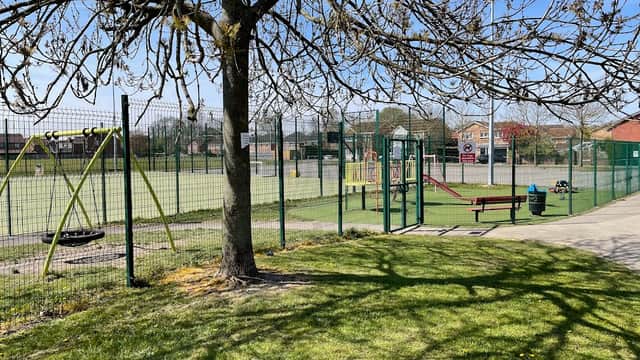 Playground with swings and climbing frame, surrounded by a fenced sports field and trees under a clear blue sky.
