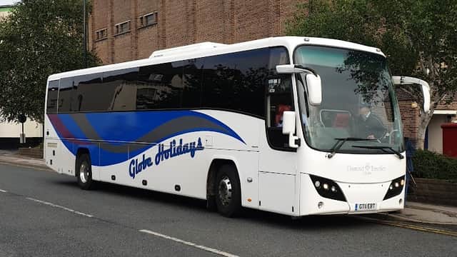 A large white coach with blue accents parked on a street near a brick building, surrounded by trees.