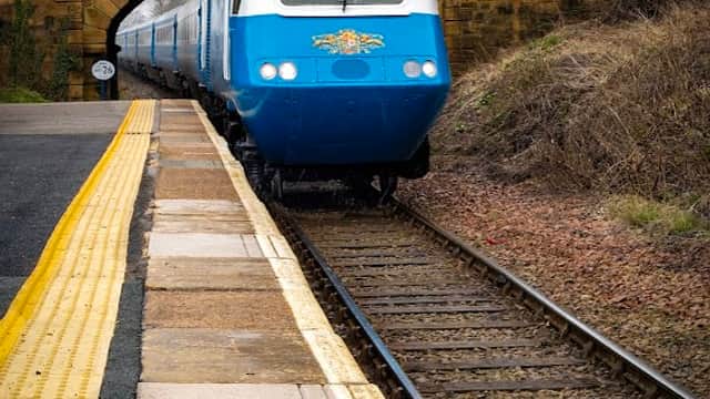 A blue train approaches a stone bridge at a railway station with a yellow platform edge and trees in the background.
