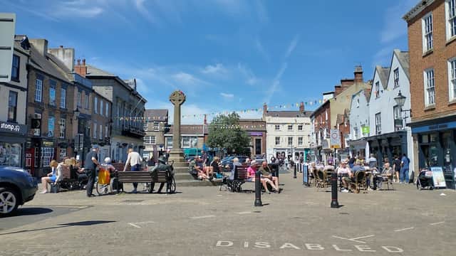 Bustling market square in Knaresborough with benches, people socializing, shops, and colorful bunting against a blue sky.