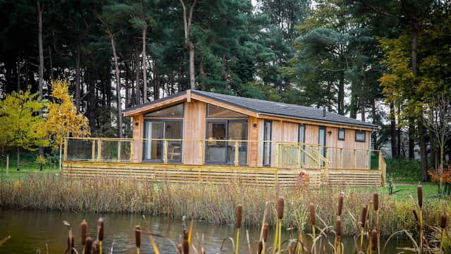 Modern wooden lodge with large windows, surrounded by trees and a pond, featuring a deck and autumn foliage.