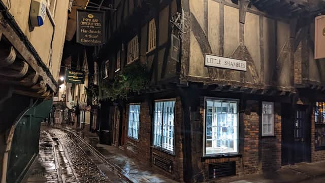 Historic narrow alley with timber-framed buildings, signage for "Little Shambles," and shop windows reflecting light.