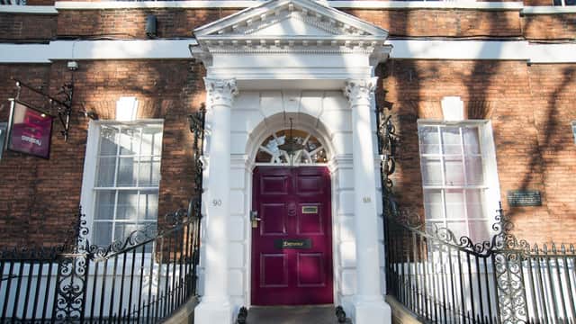 Historic brick building with a grand white entrance and a maroon door, framed by decorative iron railings.
