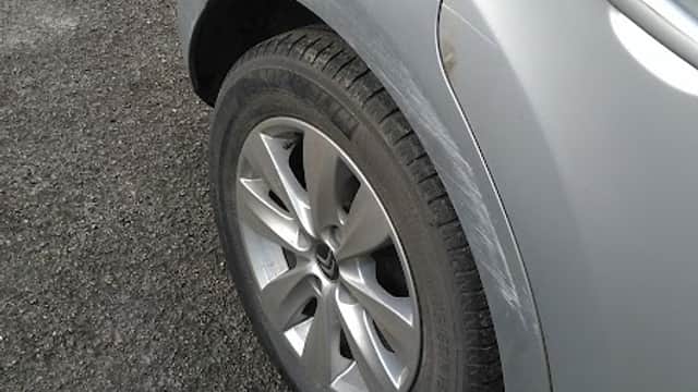 Silver car with a damaged rear wheel and tire on a gravel surface outside a body repair shop.