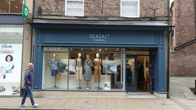 Seasalt Cornwall storefront displaying clothing and accessories, with a pedestrian walking by on a cobblestone street.