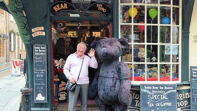 A man waves beside a large teddy bear outside a charming toy shop with colorful balloons and signs in York.