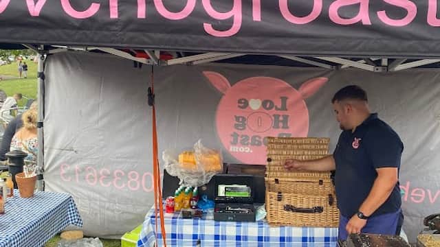 Market stall with a black canopy, blue checkered tablecloth, and a vendor preparing food. Sign reads "Love Hog Roast."