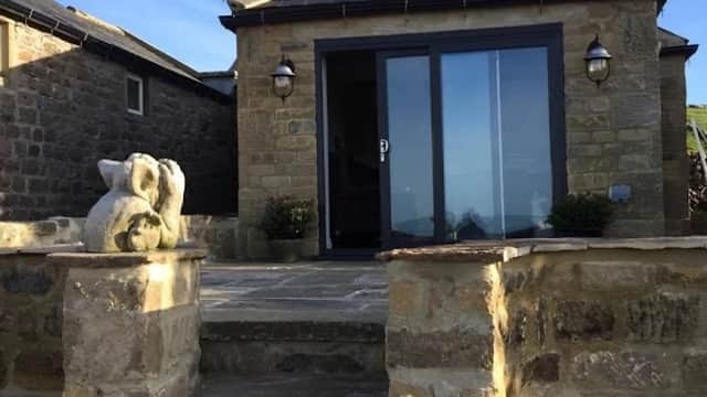 Stone cottage entrance with large glass doors, lanterns, and a stone staircase, set against a clear blue sky.