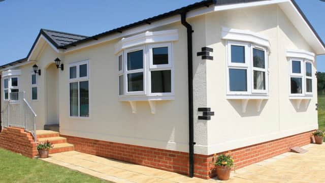 Modern mobile home with white exterior, multiple windows, and a brick patio surrounded by green grass.