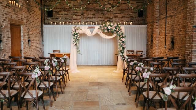 Floral-decorated wedding venue with wooden chairs, a floral arch, and soft drapery against a rustic brick backdrop.