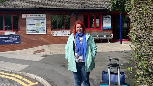 A woman with red hair stands near a blue suitcase outside Northallerton Station, with a sign and benches visible.