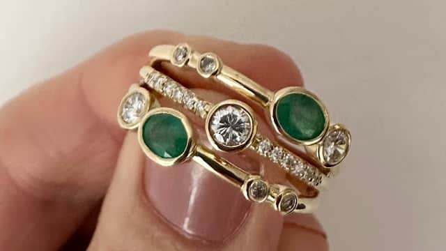Gold rings featuring emeralds and diamonds, stacked together, held between fingers against a white background.