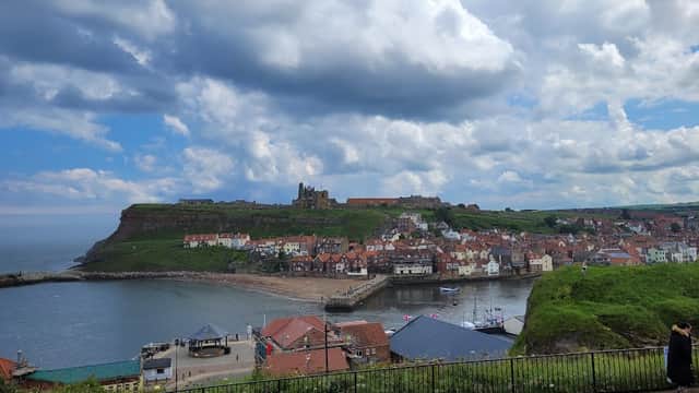 Panoramic view of Whitby with colorful buildings, a harbor, and cliffs under a partly cloudy sky.