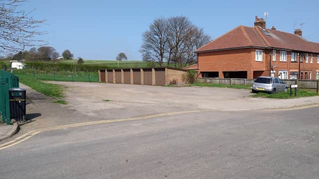 Pay & Display car park with garages, a residential building, and green fields in the background under a clear blue sky.