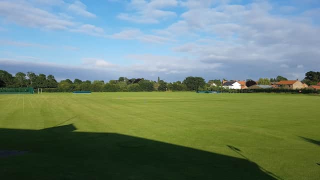 Green grass field with a cloudy blue sky, surrounded by trees and houses in Bishop Monkton, Yorkshire.